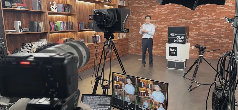 서재 세트에서 프롬프터 카메라로 강연을 촬영하며 모니터로 확인하는 장면