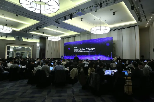 2026 HMG 글로벌 IT 포럼 행사장 — 대형 LED 화면 앞 원형 테이블에 노트북을 펴고 앉은 참석자들
